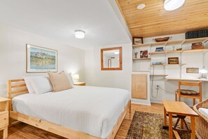 Een slaapkamer met een houten bed, een nachtkastje, een spiegel, een plank met boeken en decoratieve objecten, en een bureau met een stoel.