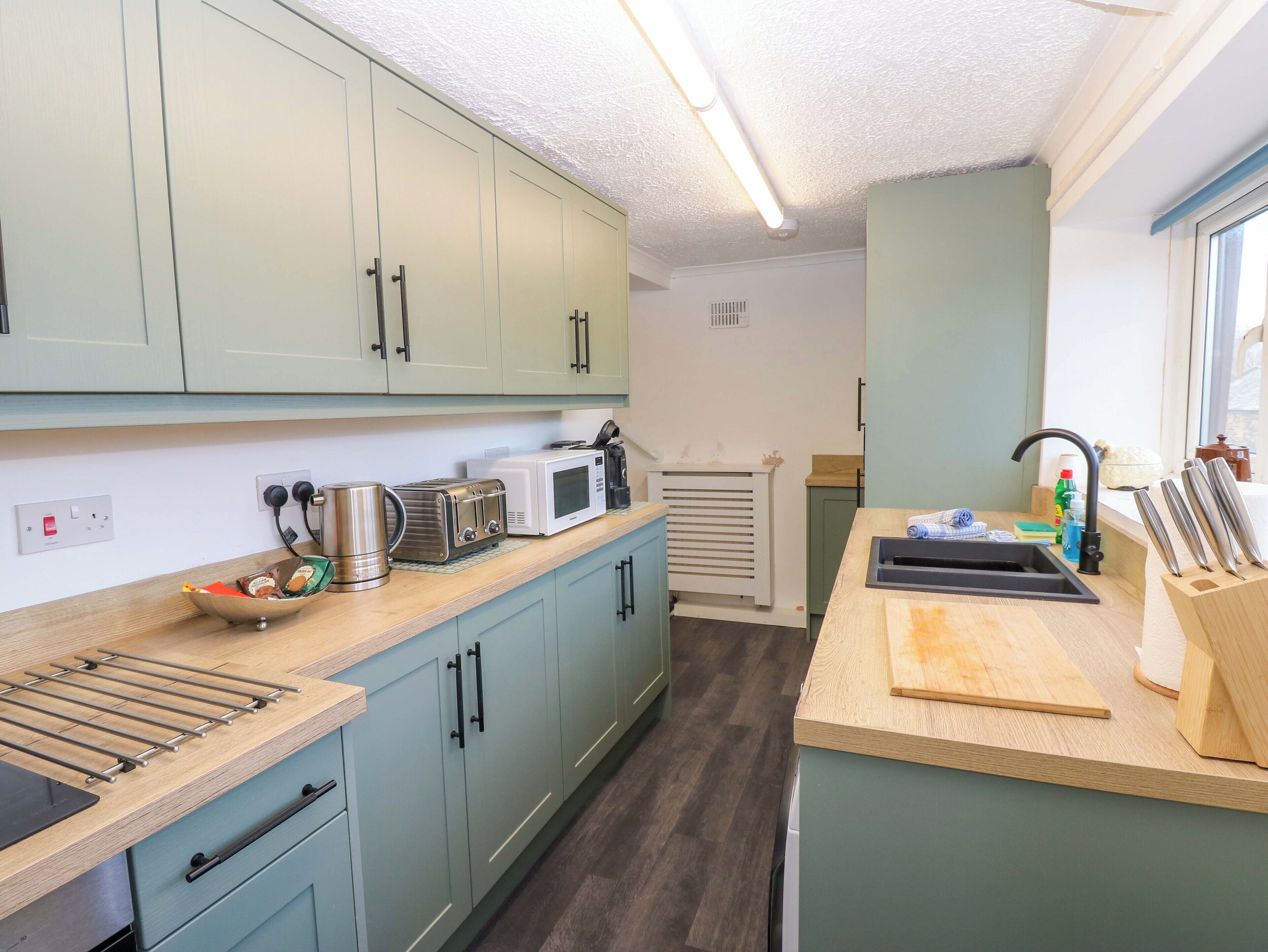 A compact kitchen with light green cabinets, a microwave, toaster, kettle, and a sink with a cutting board.