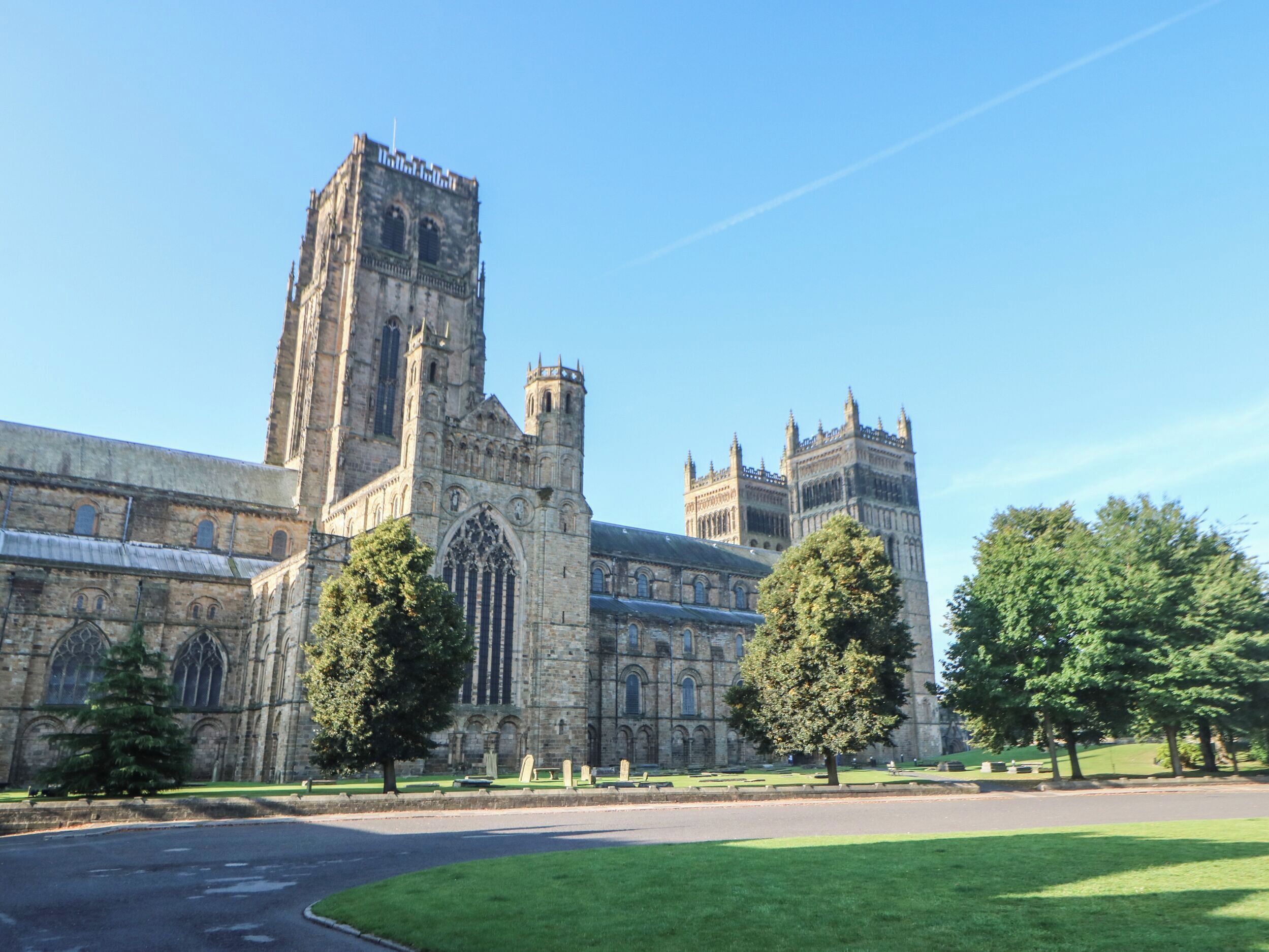 A historic cathedral with a tall central tower and two smaller towers, surrounded by a well-maintained lawn and a paved road.