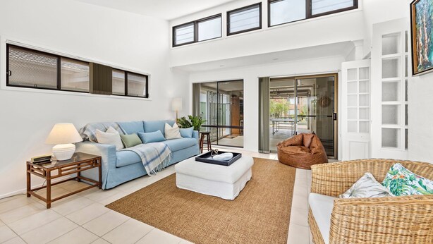 A spacious living area with a blue sofa, a white ottoman, a wicker chair, and a glass door leading to an outdoor patio. - Bay Bliss - Fireplace - 3 Mins Walk to Beach (Callala Beach)
