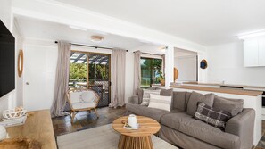 Uma sala de estar moderna com um sofá cinza, uma poltrona de vime, uma mesa de centro redonda de madeira e uma área de cozinha com armários brancos.