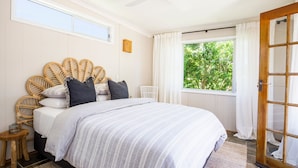 Une chambre à coucher avec un lit, une tête de lit en osier, des rideaux blancs et une vue sur la verdure à l’extérieur.