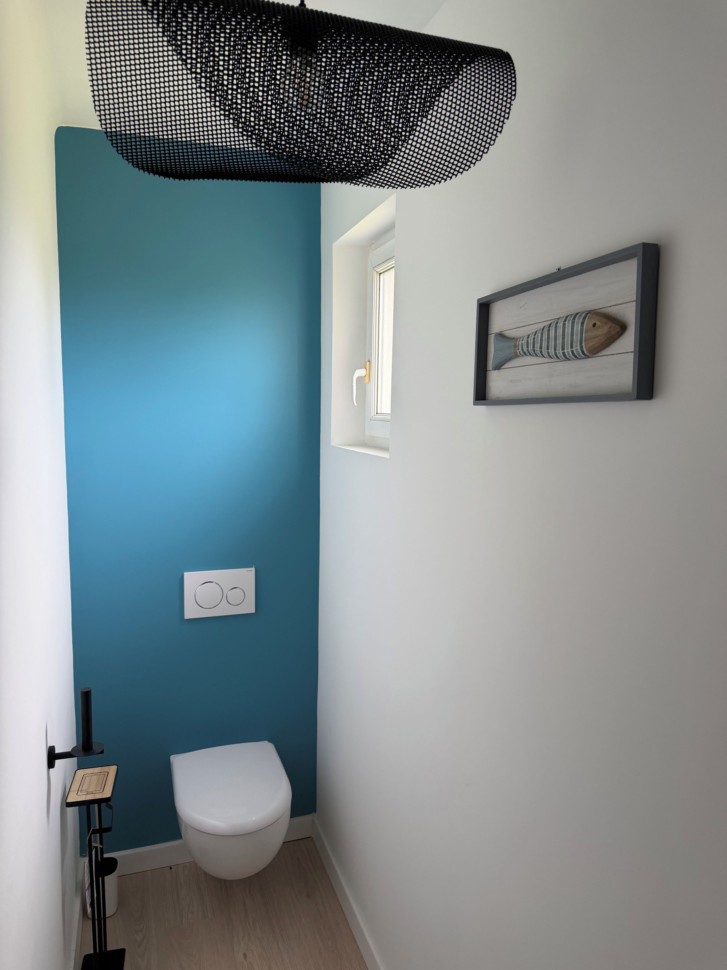 Un baño con pared azul, inodoro blanco, una obra de arte de peces en marco y lámpara de malla negra.