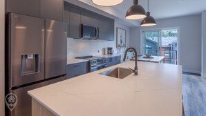 Une cuisine moderne dotée d’appareils électroménagers en acier inoxydable, d’un grand îlot central avec un évier, d’un éclairage suspendu et offrant une vue sur l’extérieur.
