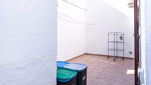 En smal utomhuskorridor med ett klinkergolv, en hylla av metall och tre soptunnor.