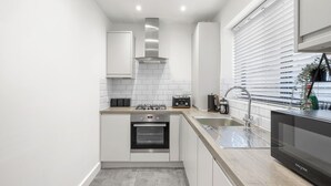 A modern kitchen with white cabinets, a stainless steel range hood, a built-in oven, a dishwasher, and a window with blinds. - Cozy  2-bedroom apartment in Canning Town near Excel London (London)