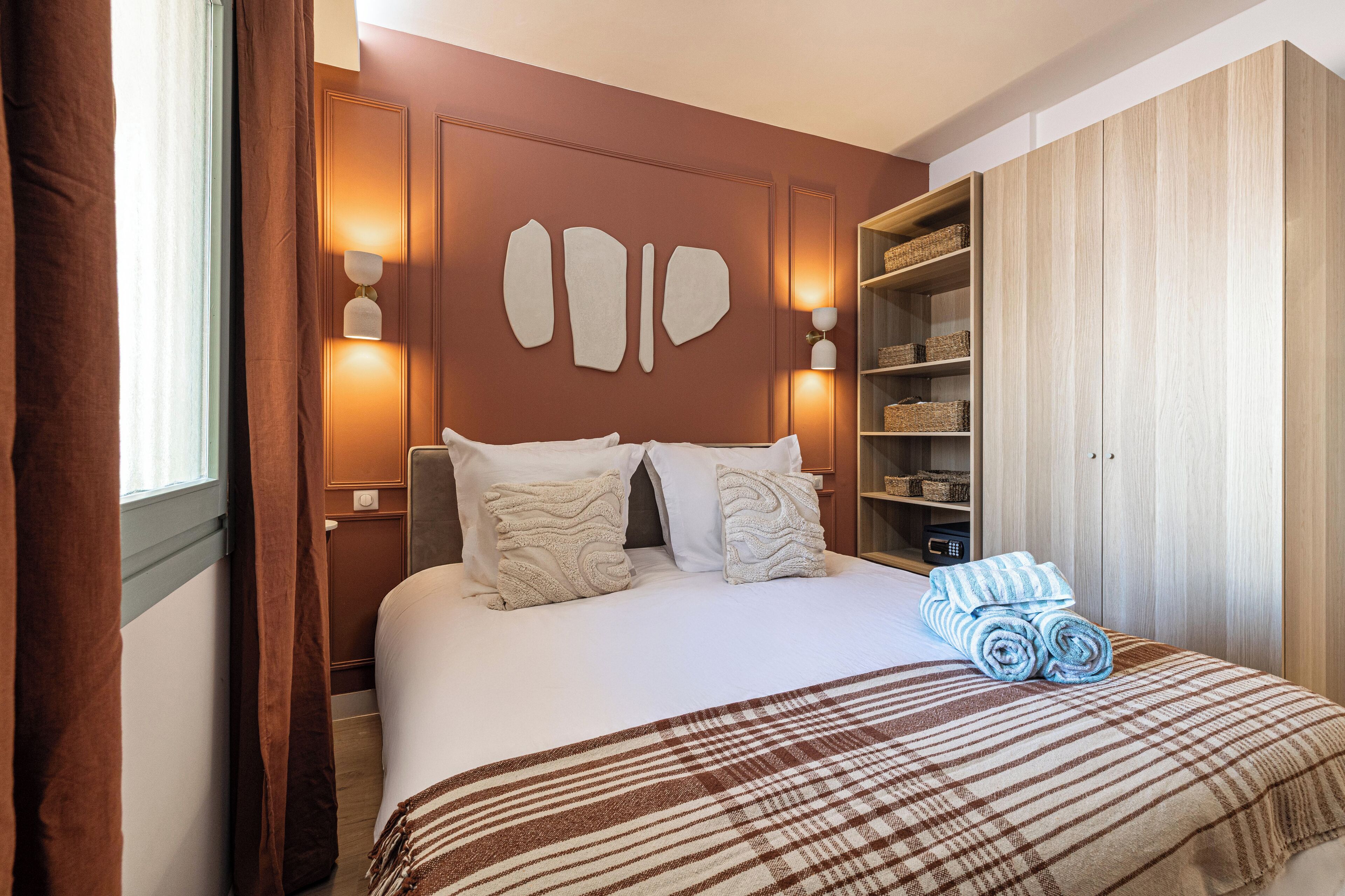 Un lit bien fait, avec une couverture à carreaux, deux oreillers et des serviettes enroulées, le tout posé sur une tête de lit en bois.