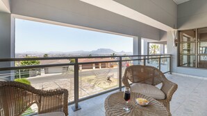 Ein Balkon mit Korbmöbeln, einer Glastrennwand und Blick auf eine Stadtlandschaft und Berge.