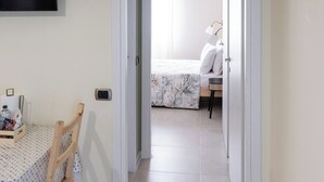 Un couloir mène à une chambre à coucher qui contient un lit, une table de chevet et une lampe. Un téléviseur est fixé au mur, et une table avec des chaises se trouve au premier plan.