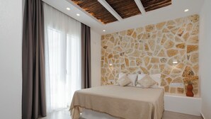 A bedroom with a stone wall, a bed with a beige bedspread, a window with curtains, and a ceiling with recessed lighting. - Villa THÉA II, Villa THÉA Naxos (Vivlos)