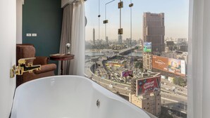 Une chambre d’hôtel moderne dotée d’une grande baignoire, d’un fauteuil en cuir, d’une petite table avec un vase, et offrant une vue sur le paysage urbain, avec un pont et des immeubles.