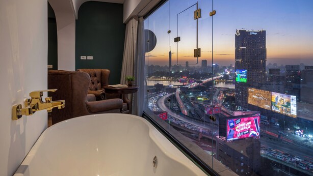 Une chambre d’hôtel moderne dotée d’une grande baignoire, d’un fauteuil en cuir et offrant une vue sur le paysage urbain au crépuscule.