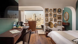 Une chambre d’hôtel avec vue sur la ville, un bureau avec une chaise, un lit et un mur décoré de tableaux encadrés et d’un gramophone.
