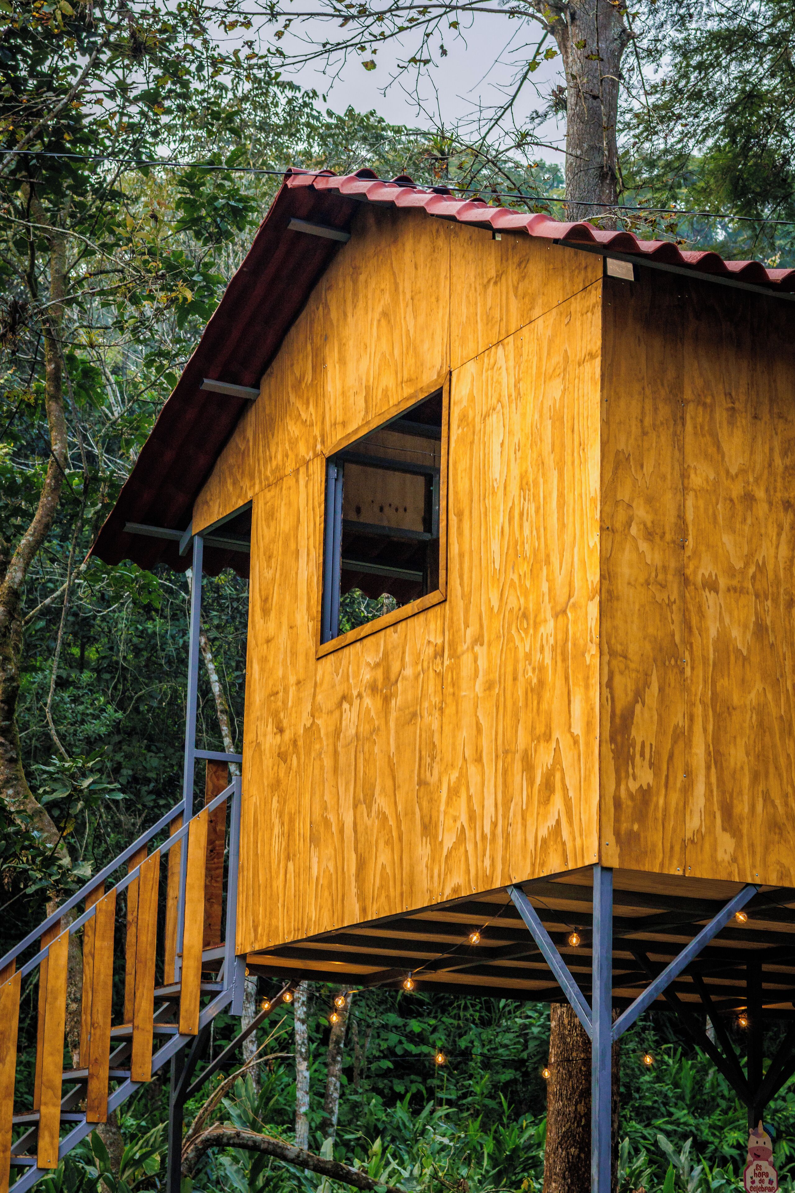 A wooden treehouse with a red roof, built on stilts, and surrounded by lush greenery.