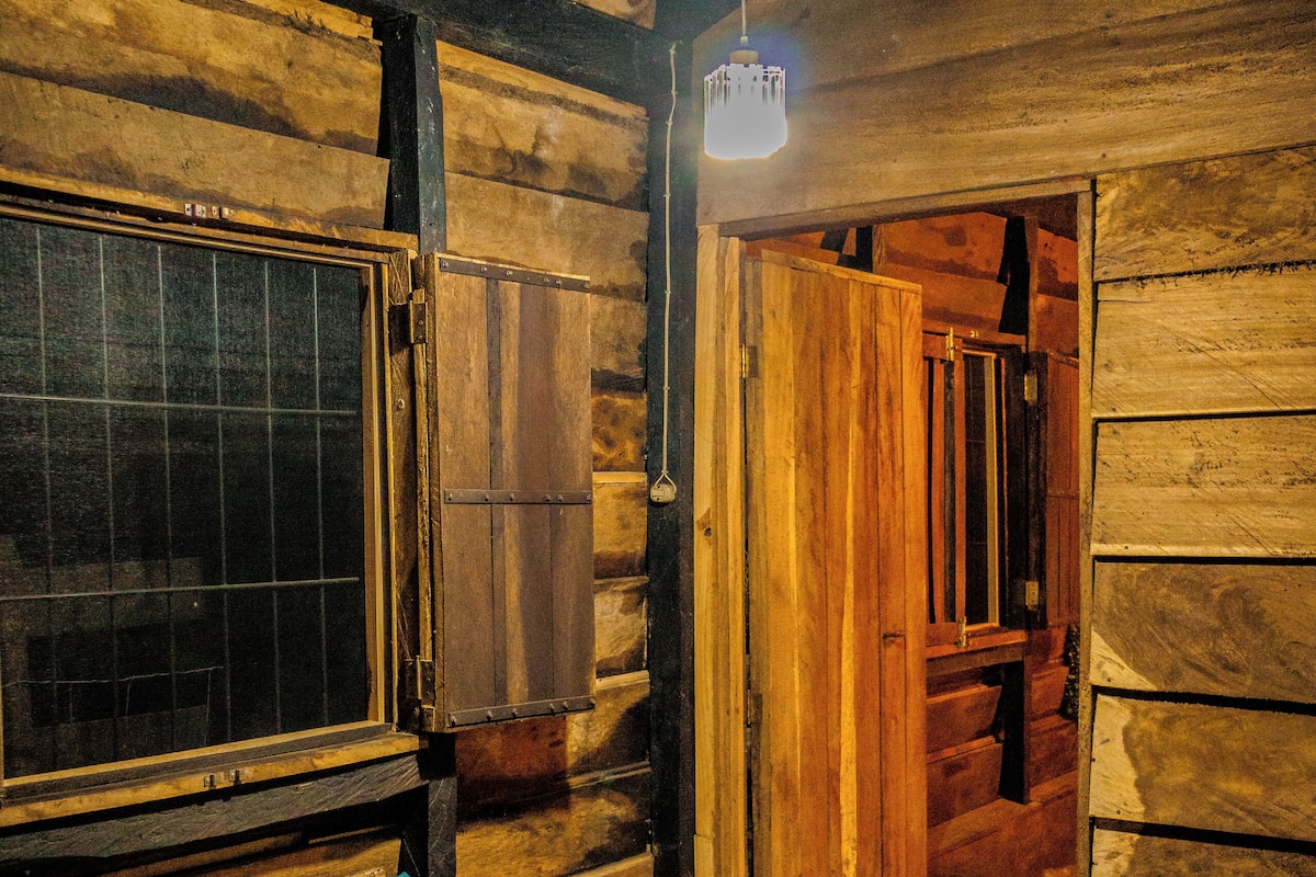 Interior rústico de una cabaña de madera con una silla de mimbre, una alfombra morada y una lámpara colgante.