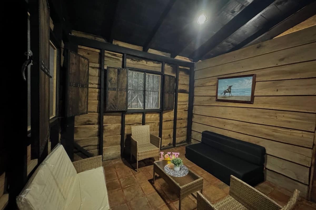 Una habitación acogedora con paredes de madera y un ventanal grande. Hay un sofá, una mesita con una planta y dos sillones de mimbre.