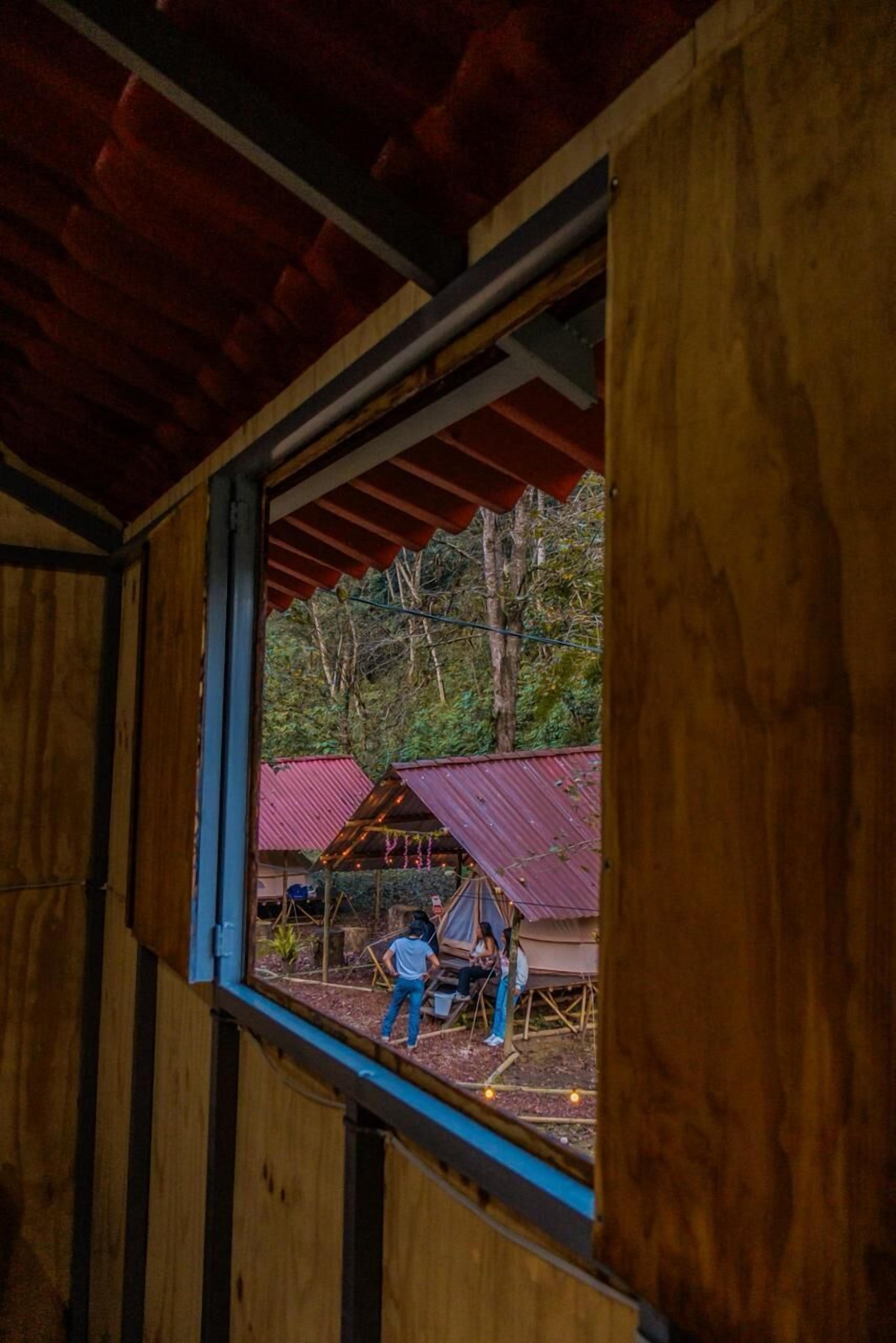 Un interior de madera con un ventanal que muestra un espacio exterior con construcciones de techo rojo y árboles.