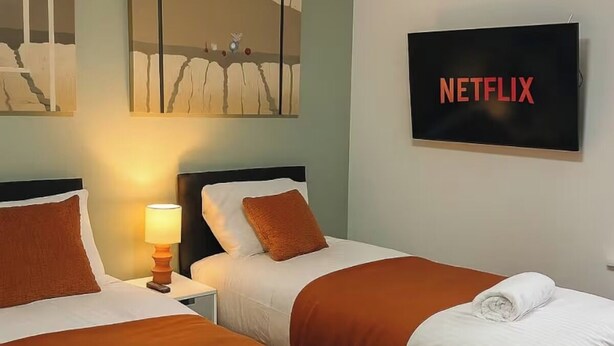 Een hotelkamer met twee bedden, een nachtkastje met een lamp, een wandtelevisie waarop 'NETFLIX' wordt weergegeven, en een groot abstract schilderij boven de bedden.