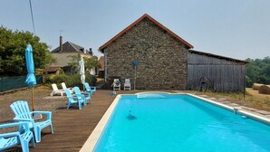 Ein Swimmingpool mit Holzterrasse, blauen Stühlen und einem Steinhaus im Hintergrund.