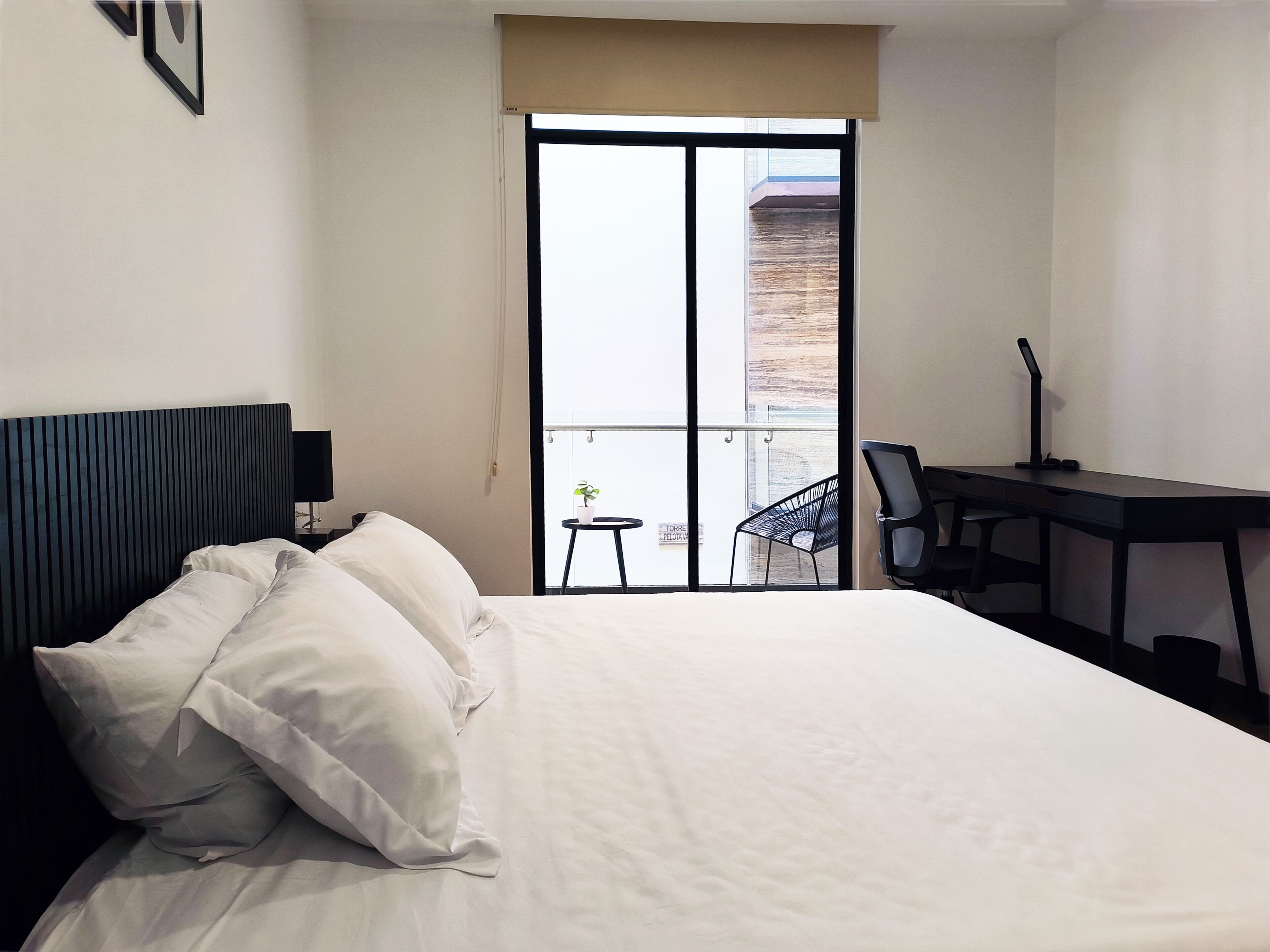 A hotel room with a bed, a desk with a chair, a window, and a balcony with a plant.
