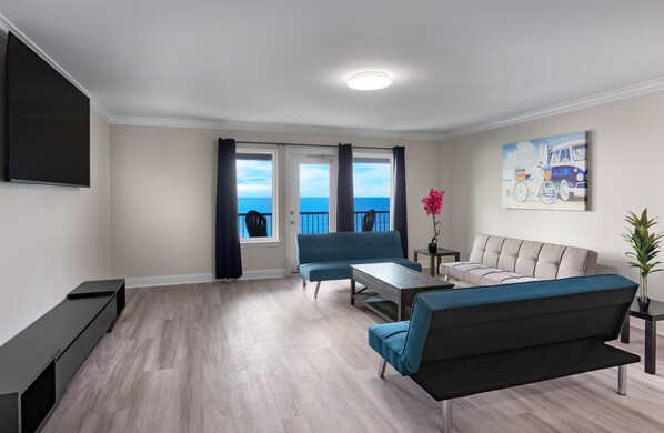 A modern living room with a sofa, a coffee table, and a TV. There are large windows with curtains, a painting on the wall, and a potted plant. - New Snowbird 3BR Condo On The Boardwalk + 100 Dollar Resort credit (Myrtle Beach)