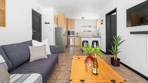 Een moderne woonkamer met een bank, een houten salontafel met een fles wijn en een glas, een keuken op de achtergrond, en een aan de muur bevestigde televisie.