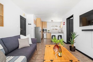 A modern living room with a sofa, a wooden coffee table with a wine bottle, and a kitchen in the background. - Big Bay Beach Club 182 (Cape Town)