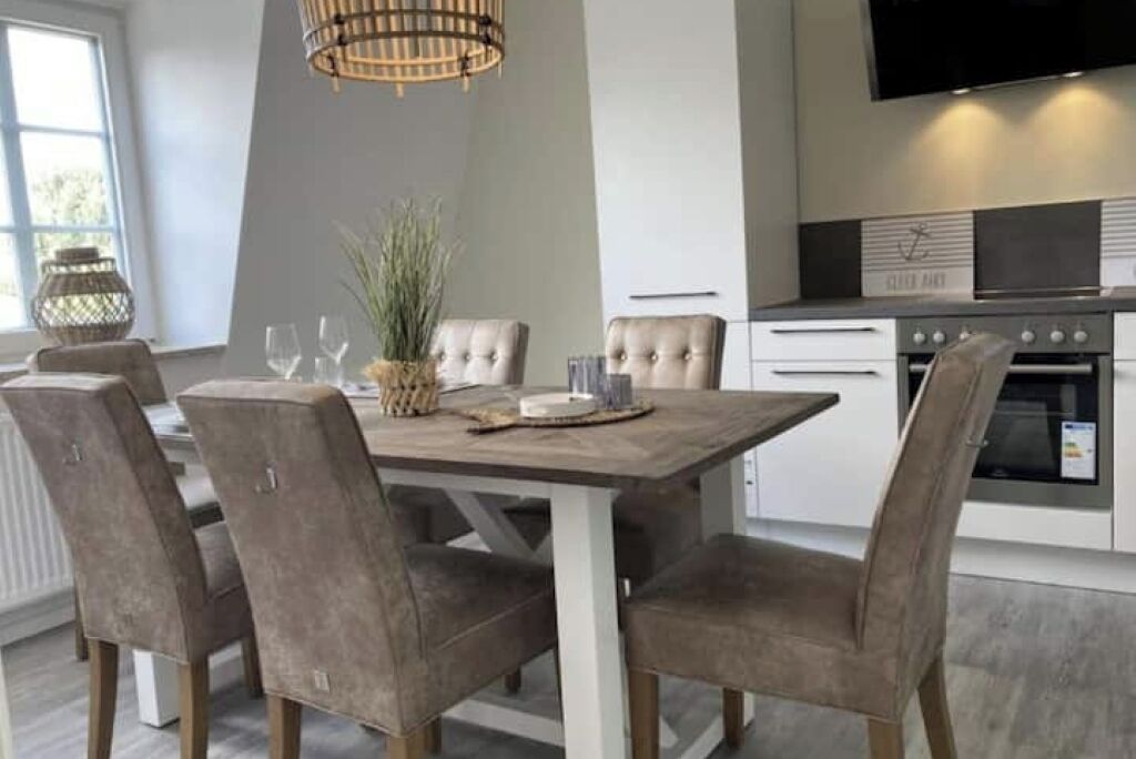 A modern kitchen with a wooden dining table, four upholstered chairs, a hanging light fixture, and built-in appliances.