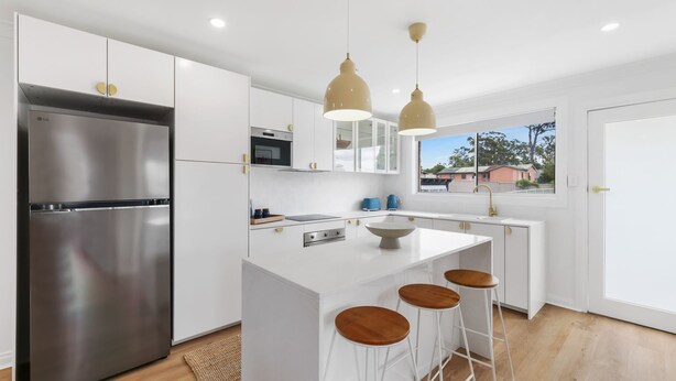 A modern kitchen with white cabinetry, a stainless steel refrigerator, a central island with stools, pendant lighting, and a window with a view of houses. - The Pearl by Book Jervis Bay A Fresh, Modern Family Retreat in Old Erowal Bay (Old Erowal Bay)