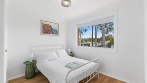 A bedroom with a bed, a bedside table, a lamp, a framed picture, and a window with a view of trees and buildings. - The Pearl by Book Jervis Bay A Fresh, Modern Family Retreat in Old Erowal Bay (Old Erowal Bay)