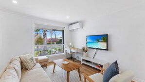 A modern living room with a large window, a flat-screen TV, a sofa, a coffee table, and a desk. - The Pearl by Book Jervis Bay A Fresh, Modern Family Retreat in Old Erowal Bay (Old Erowal Bay)