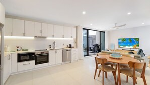 A modern kitchen with integrated appliances, a dining area with a wooden table and chairs, and a living space with a TV displaying a beach scene. - Husky Beach Pad by Book Jervis Bay - Walk to the beach (Huskisson)