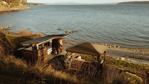 Une cabane en bois dotée d’une terrasse donnant sur un lac, entourée d’une végétation luxuriante et surplombant une chaîne de montagnes lointaine.