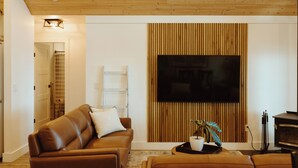 Un salon moderne avec un plafond en bois, un grand téléviseur à écran plat fixé sur un mur recouvert de panneaux en bois, un canapé en cuir marron et une petite table sur laquelle est posée une plante.