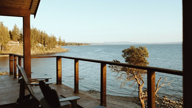 Une terrasse en bois avec des bancs, donnant sur un lac, entourée d’arbres et bordée de montagnes lointaines.