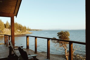 Una terraza de madera con bancos que da a un lago, rodeada de árboles y montañas a lo lejos.