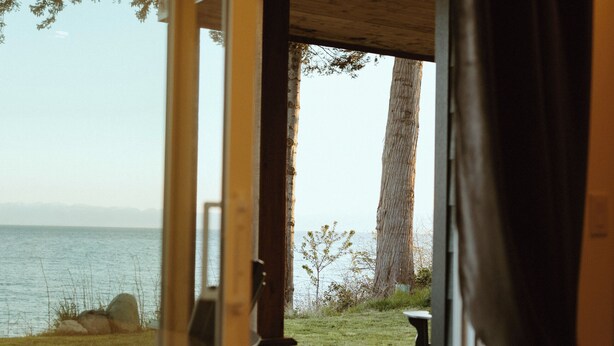 Blick von einem Zimmer mit einer Schiebeglastür zu einem Außenbereich mit Tisch und Stühlen, einem großen Baum und einem Gewässer im Hintergrund.