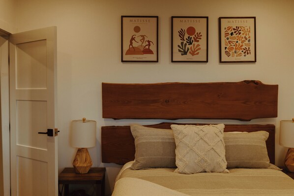 A bedroom with a wooden headboard, a bed with beige bedding, two bedside tables with lamps, and three framed artworks on the wall.