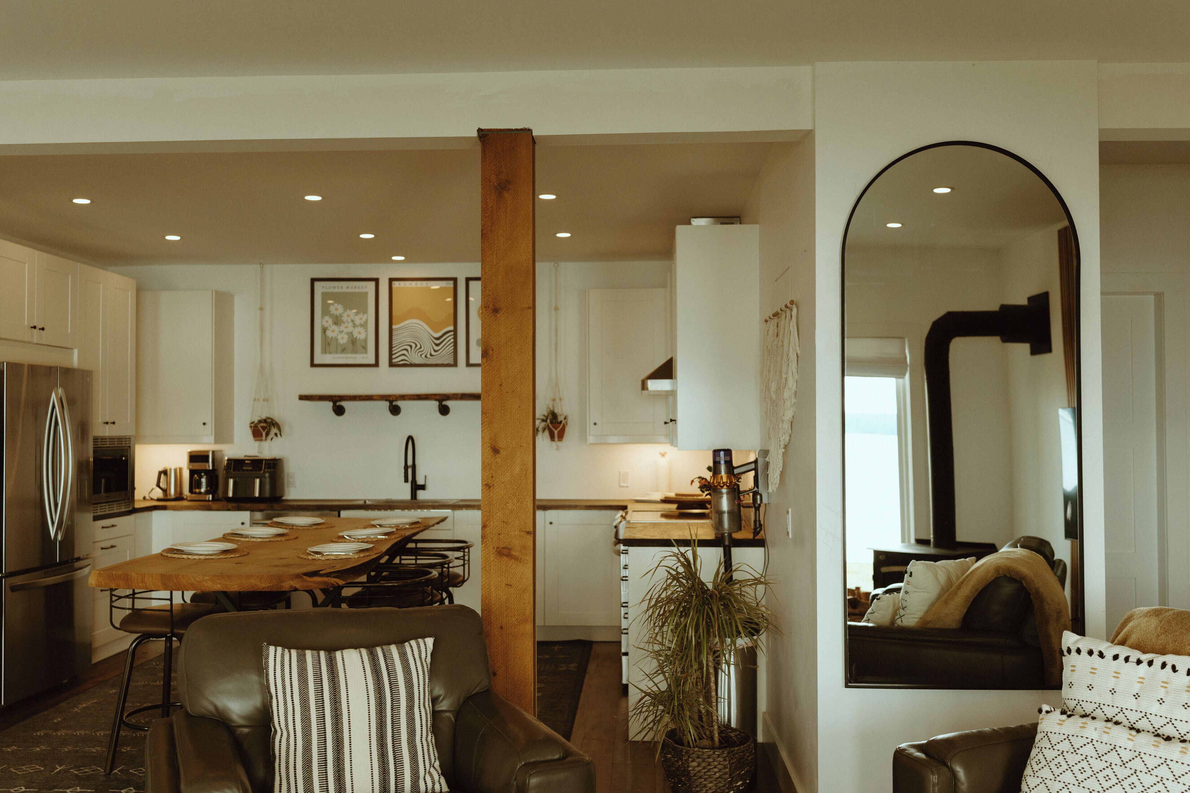 Une cuisine moderne avec des meubles blancs, une table à manger en bois et un îlot central. On y trouve également un espace salon avec un canapé en cuir et un grand miroir.