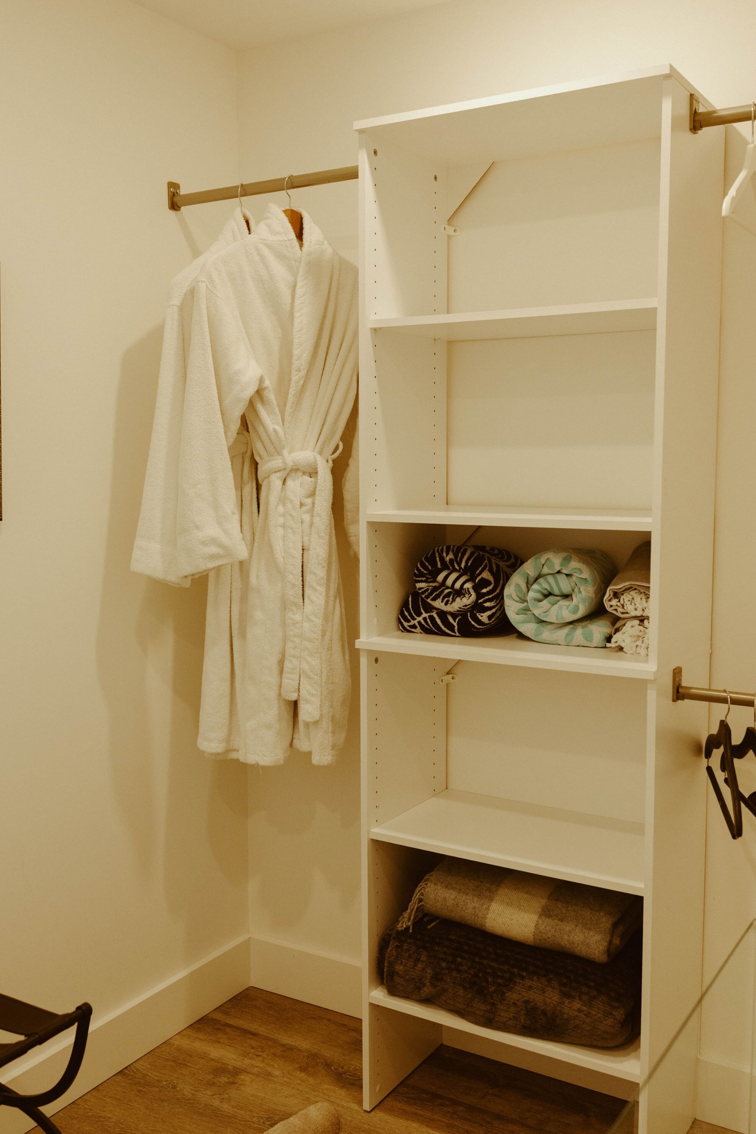 A white robe hanging on a rack, a white shelving unit with folded blankets, and a wooden floor.
