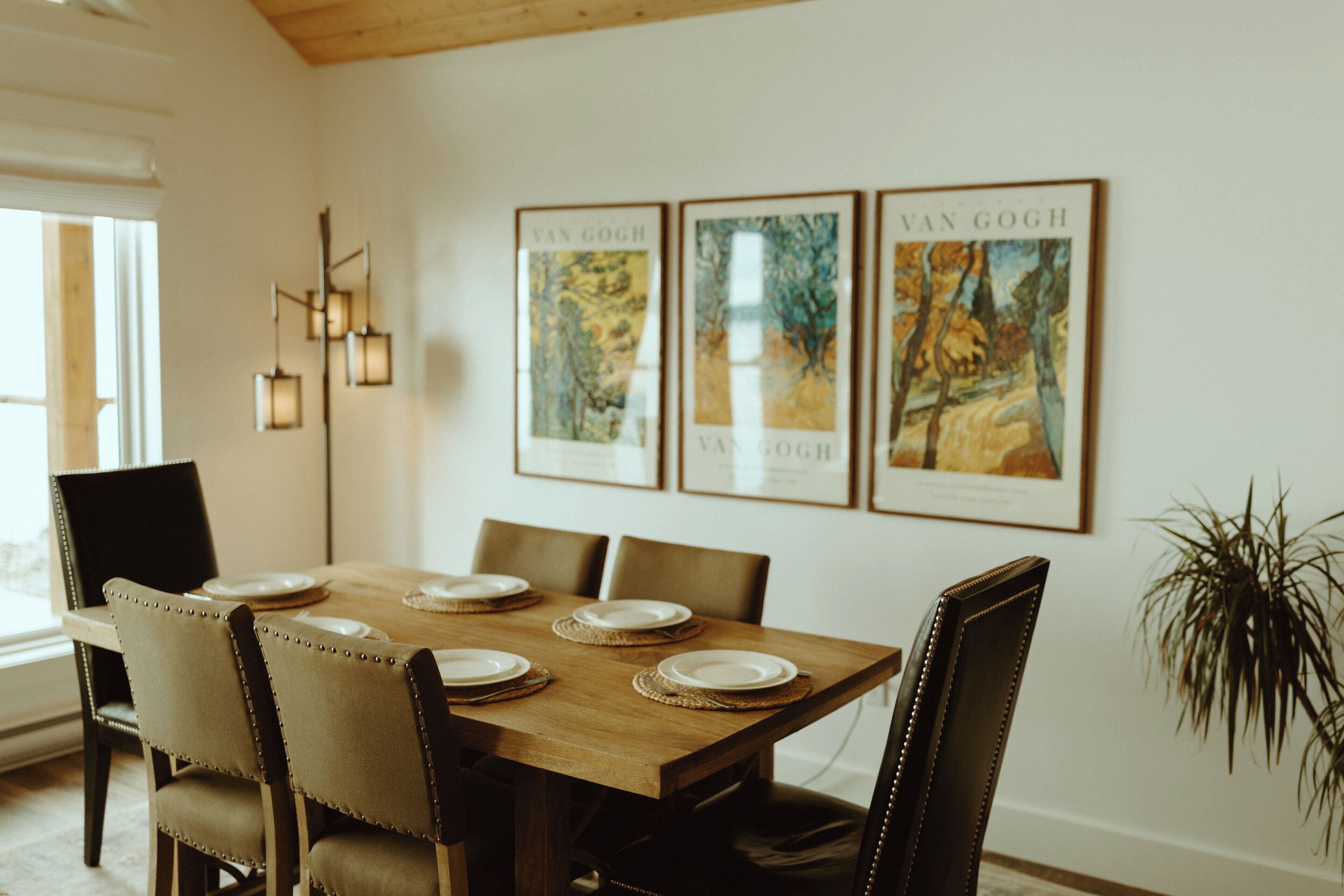 Een eetkamer met een houten tafel voor zes personen, zes stoelen en drie ingelijste Van Gogh schilderijen aan de muur.