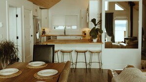 Een moderne keuken met witte kastjes, een houten eiland en barstoelen. Op de voorgrond staat een eettafel met borden en een plant.