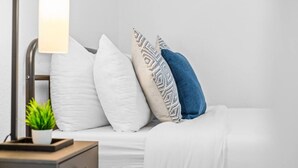 Un letto rifatto con biancheria bianca e cuscini decorati, una lampada da comodino e una piccola pianta in vaso su un tavolino.
