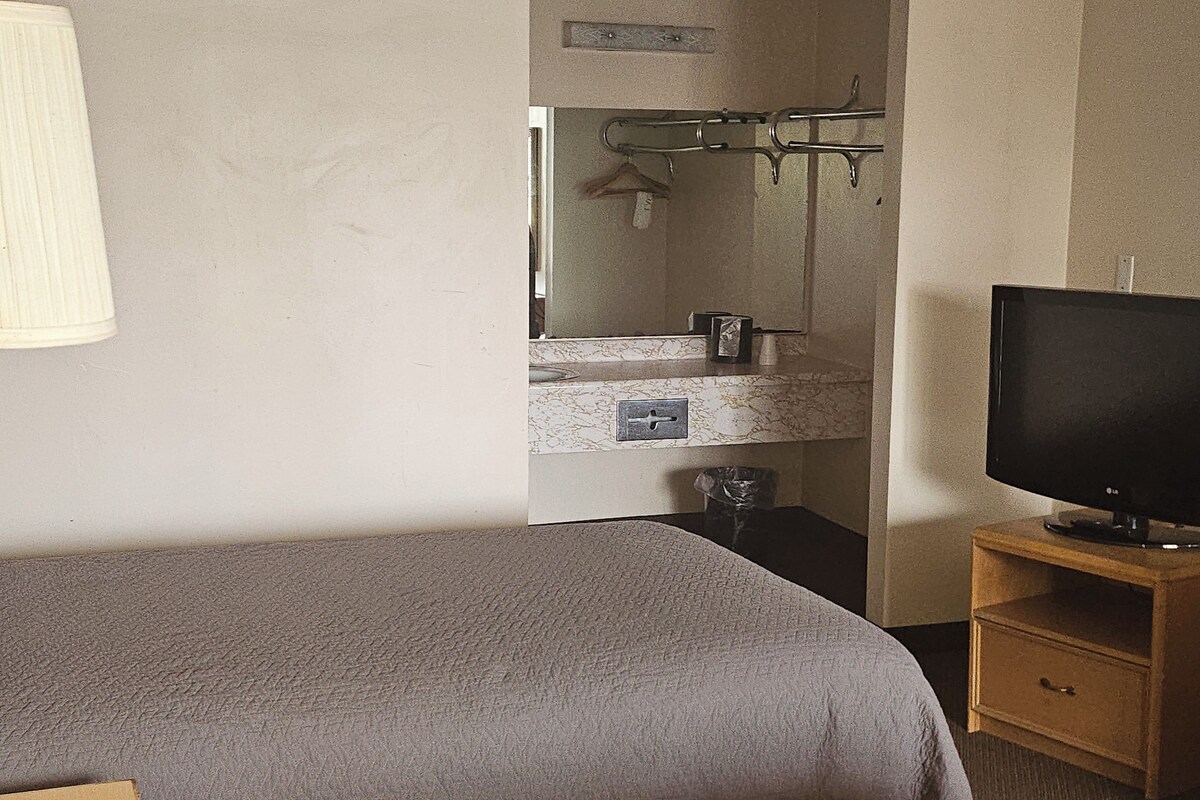 Une chambre à coucher avec un lit, un téléviseur posé sur un meuble en bois, une salle de bain avec un miroir et un porte-serviettes.