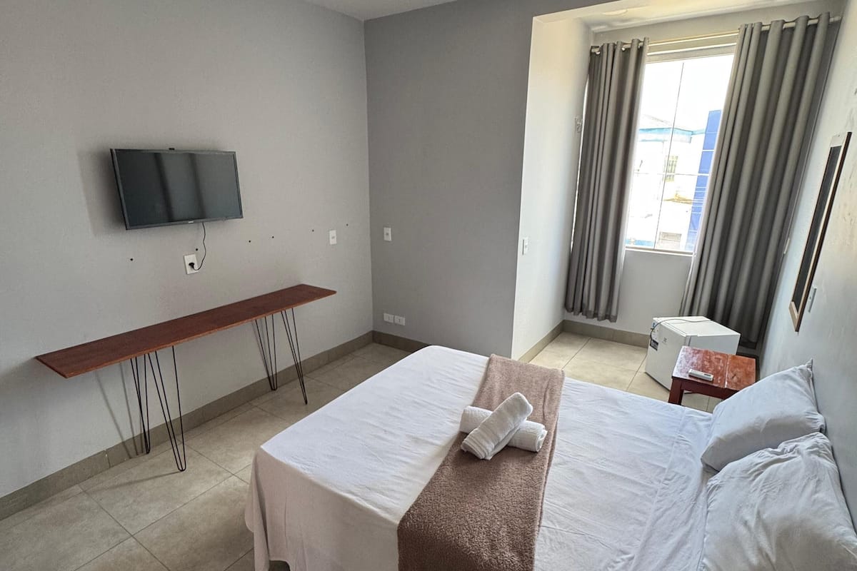 Una camera d'albergo con un letto, una scrivania, una TV a parete e una finestra con tende.