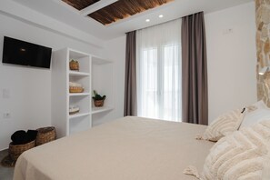 A bedroom with a large bed, a TV mounted on the wall, a shelf with baskets, and a window with curtains. - Villa THÉA III, Villa THÉA Naxos (Naxos)