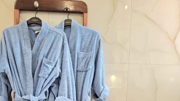 Two light blue bathrobes hanging on a wooden rack in a bathroom with marble walls and a toilet paper holder. - Tia Vahine - Boutique Hôtel Ivato (Antananarivo)