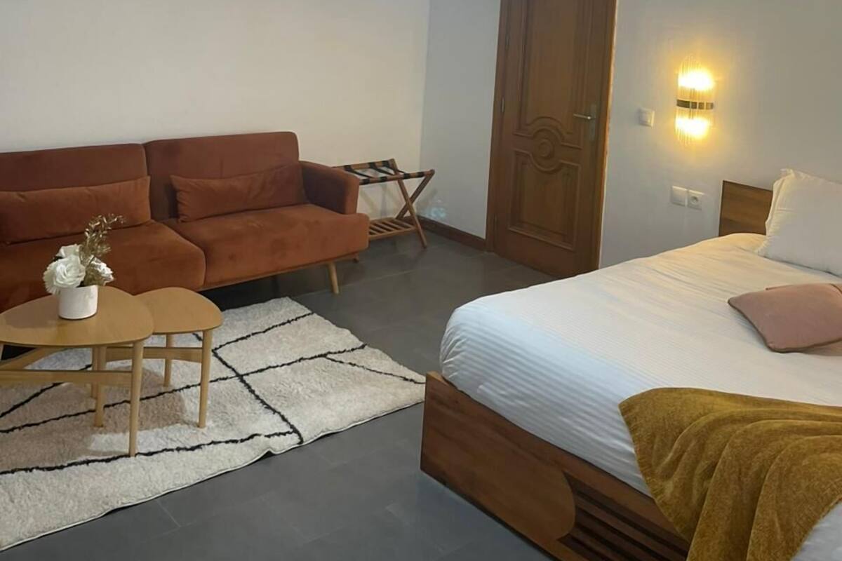 A bedroom with a bed, a wooden headboard, a brown sofa, a small round table with a vase of flowers, a white rug, a chandelier, and a wall-mounted light fixture.