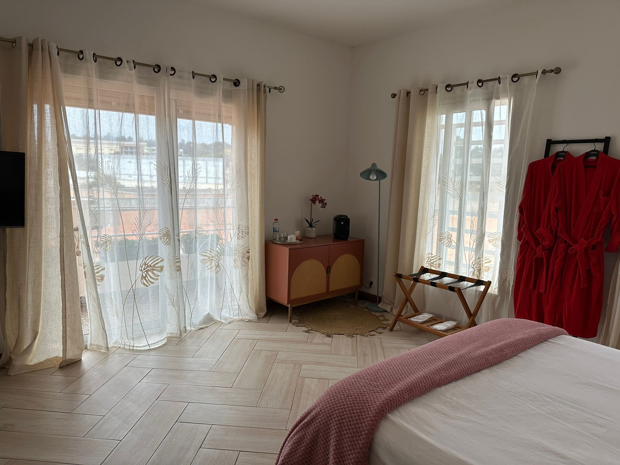Quarto com cama, cômoda, espelho, janela com cortinas leves e roupão vermelho pendurado em um suporte.