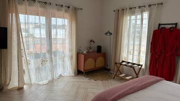 Quarto com cama, cômoda, espelho, janela com cortinas leves e roupão vermelho pendurado em um suporte.